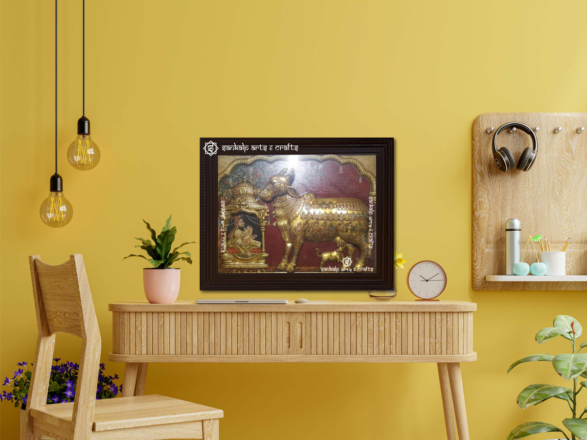 A framed Kamadhenu antique Tanjore painting displayed as a wall hanging, radiating divine energy with rich gold accents, artistic detailing, and an antique finish.